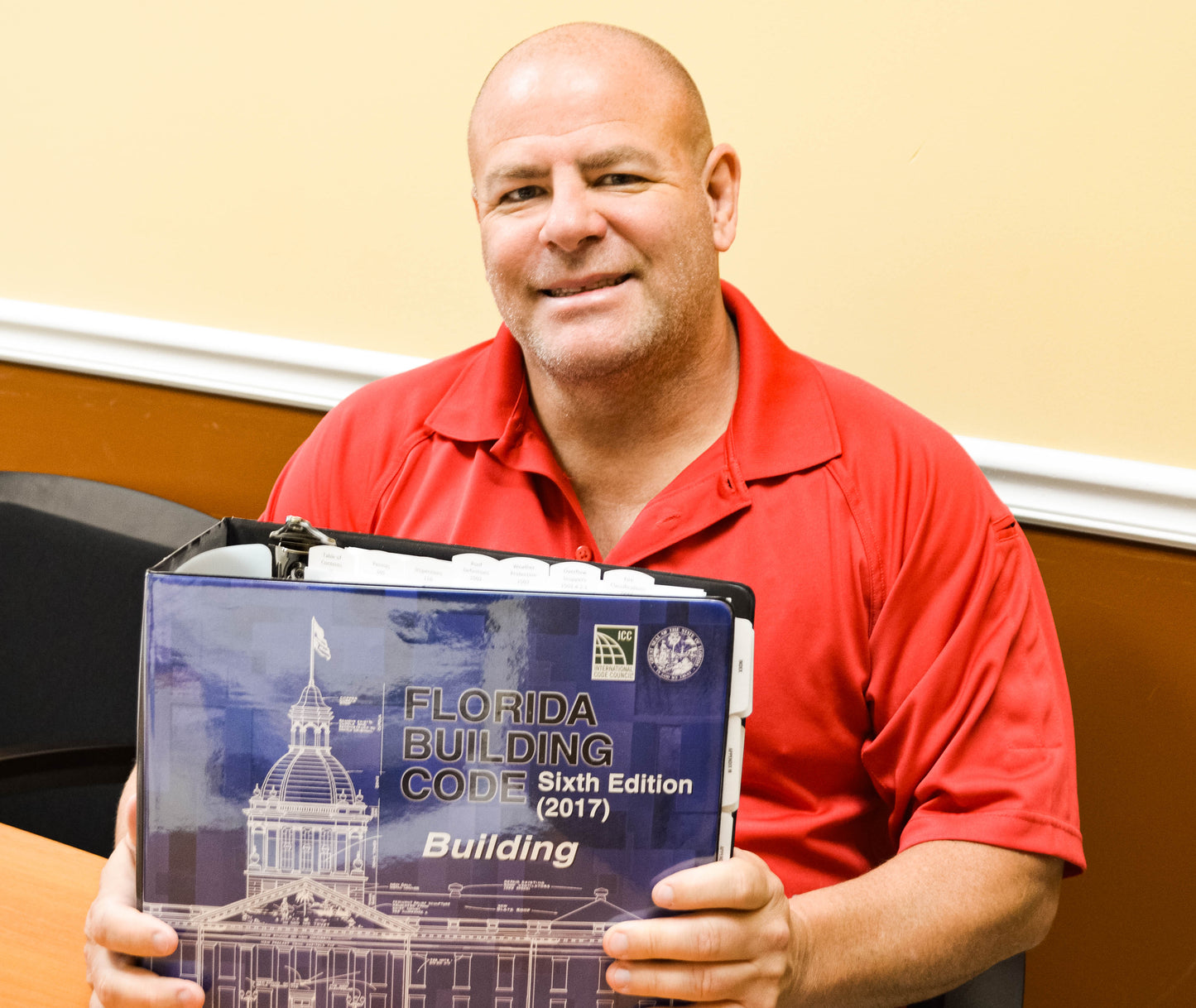 Man in red shirt holding 2017 Florida Building Code sixth edition binder with architectural drawings on cover