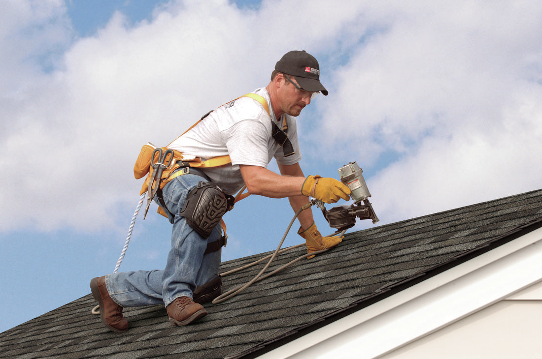 Male roofer wearing safety gear using nail gun on black shingled roof under blue sky
