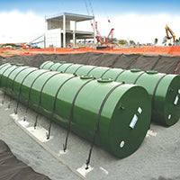 Green cylindrical underground liquid storage tanks aligned on gravel at a construction site with building structures in the background